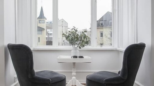 Una habitación con dos sillas negras orientadas hacia una mesa blanca y un ventanal con vista a edificios.