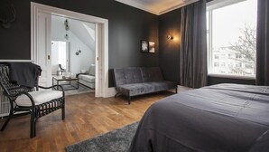 Un dormitorio moderno con una cama grande, un sofá oscuro, una silla y vistas a la ciudad a través de un amplio ventanal.
