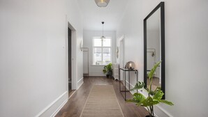 Un pasillo con suelo de madera, alfombra, planta en maceta, espejo y lámpara colgante.