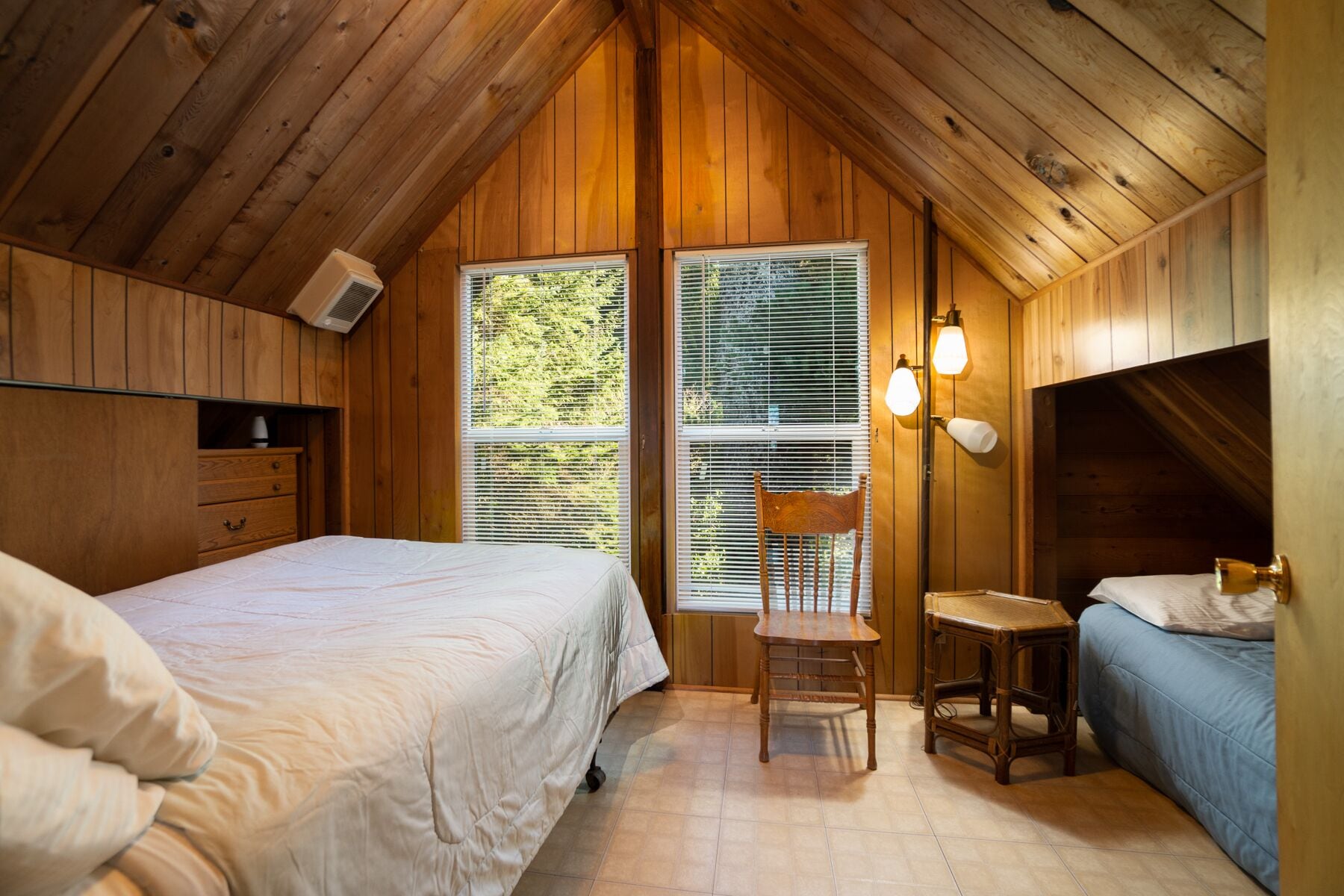A cozy cabin bedroom with two beds, a wooden ceiling, and large windows with blinds.