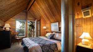 A cozy bedroom with a wooden ceiling, a large bed with patterned bedding, a desk with a lamp, and a window overlooking a tree.