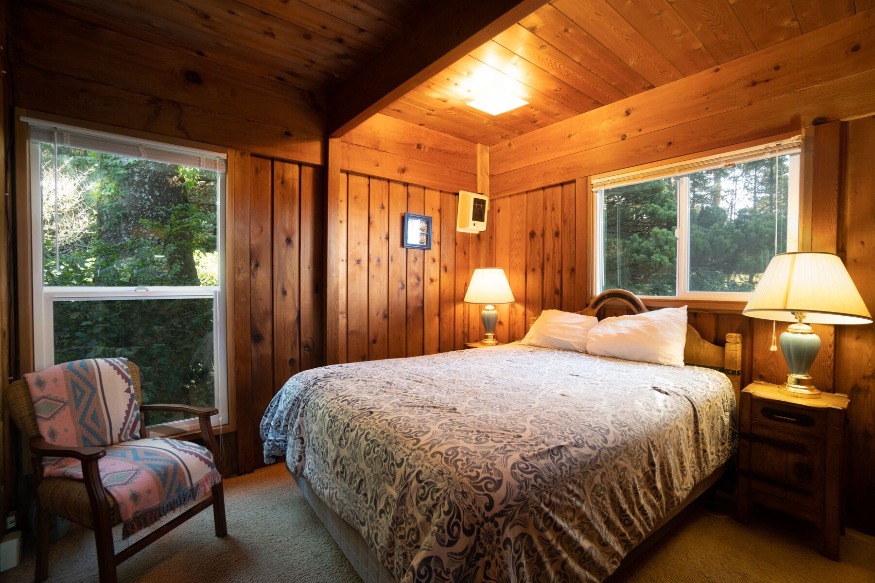 A wooden cabin bedroom with a bed, two bedside lamps, a chair, and a window.