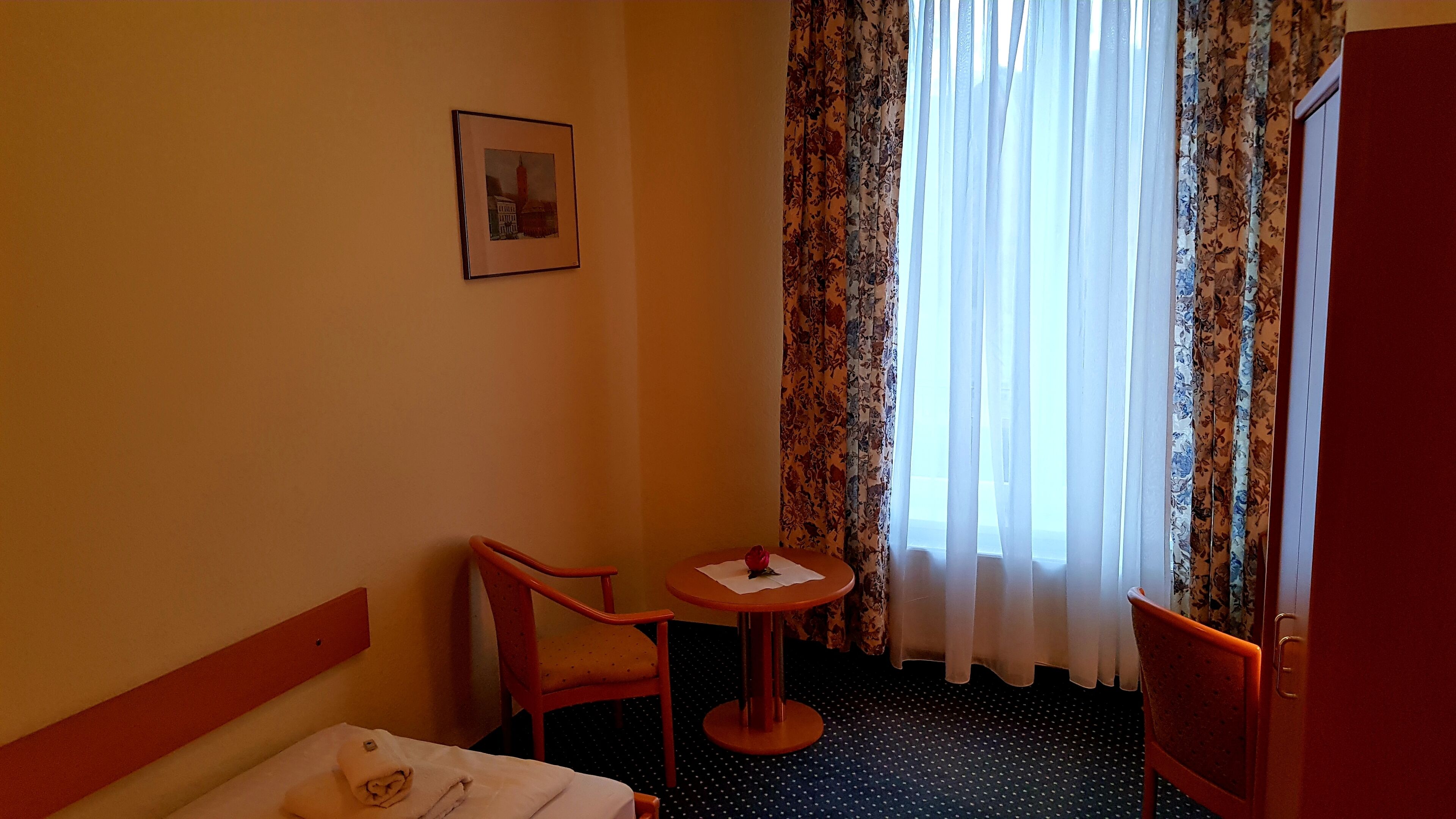 Una camera d'albergo con un letto, un tavolino con una rosa, una sedia, un quadro appeso e una finestra con tende.