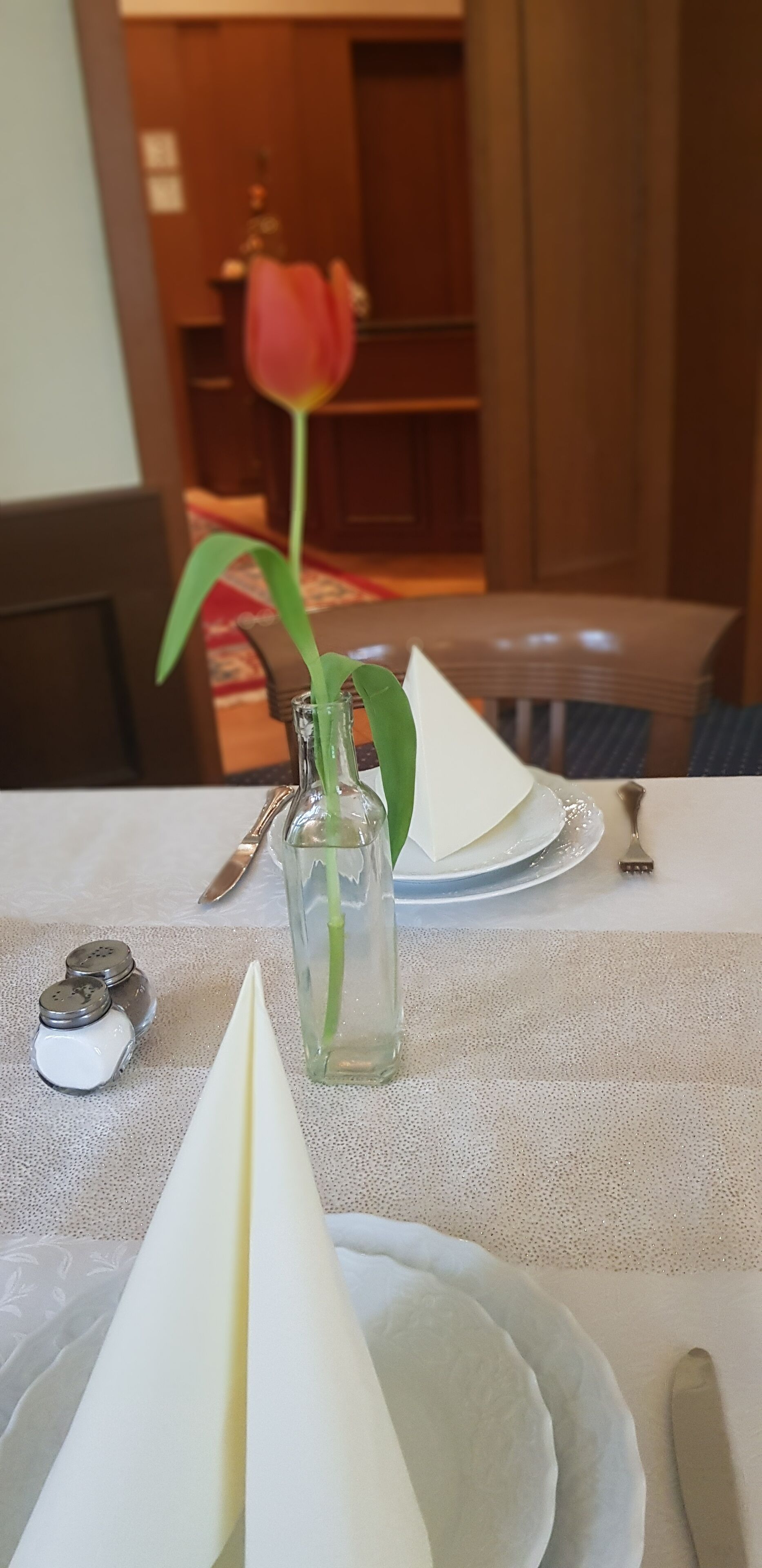 Une table à manger ornée d’un vase en verre transparent contenant une seule rose, de deux salerons et poivriers, et d’une serviette pliée.