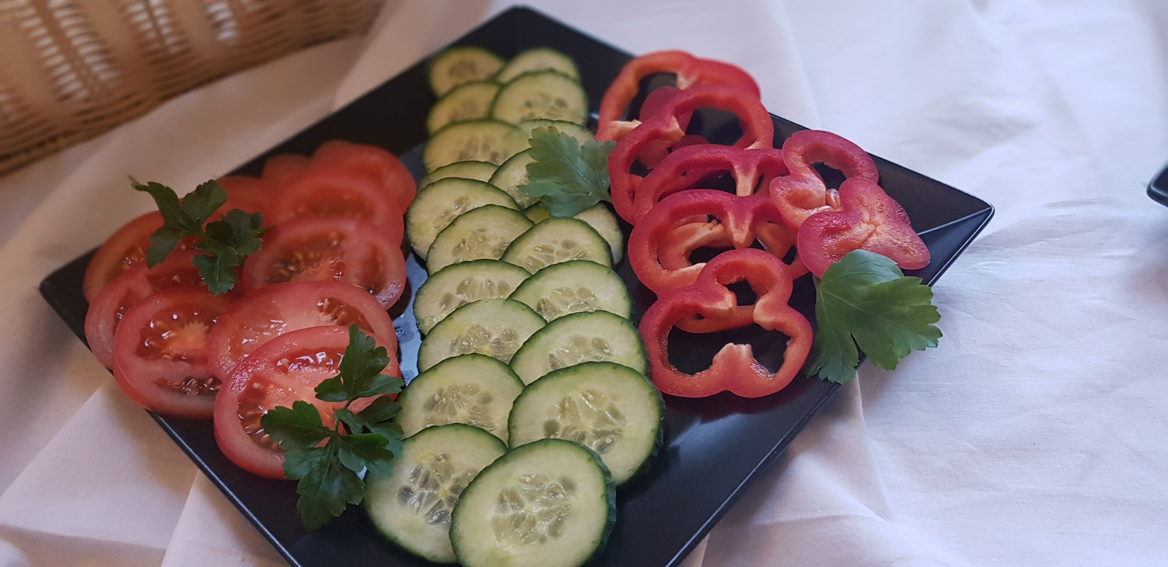 Ein schwarzer Teller mit geschnittenen Tomaten, Gurken und roten Paprika, garniert mit Petersilie, auf einer weißen Tischdecke.