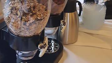 Une machine à café avec un récipient en verre rempli de granola, un pichet en métal et un bol de fruits.