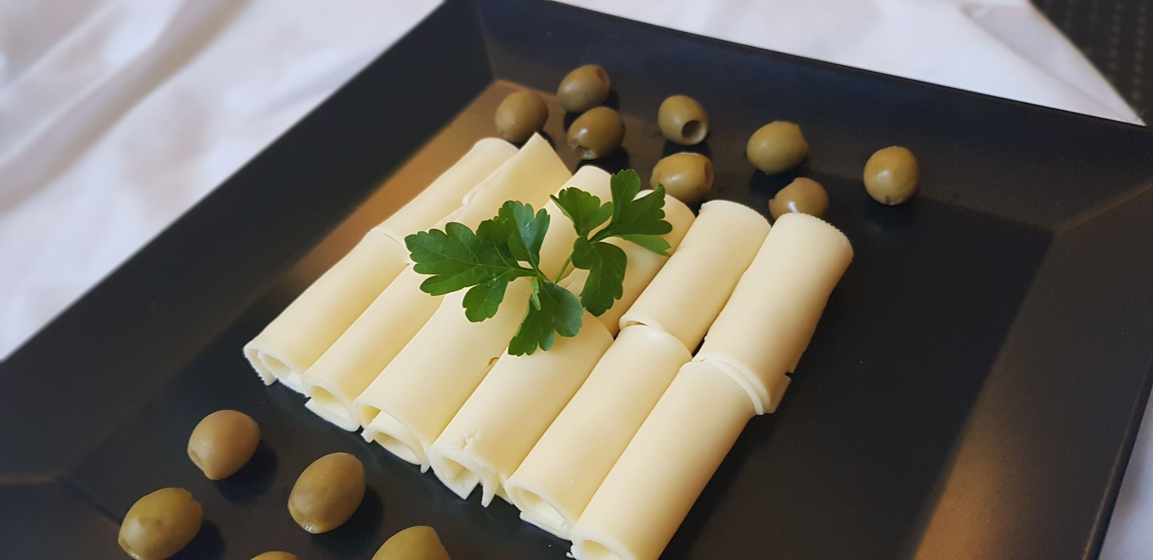 A black rectangular plate with rolled white items and green olives, garnished with a green leaf.