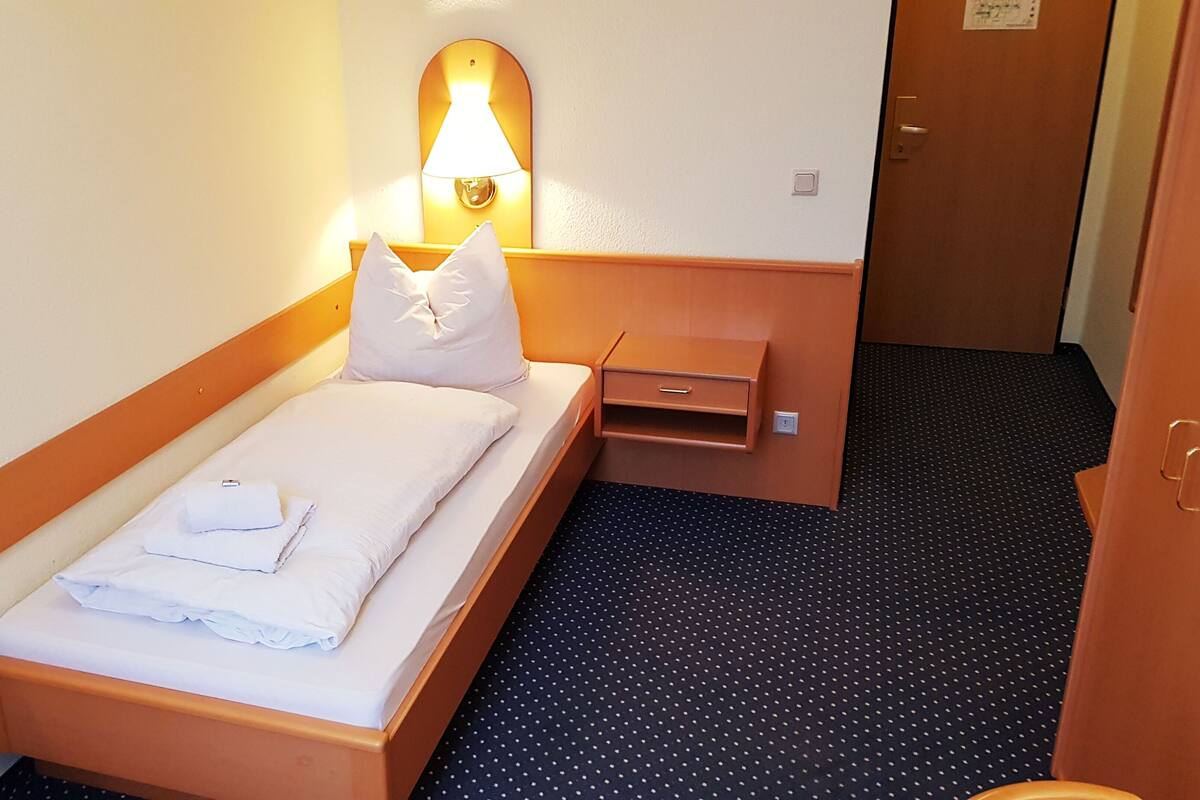 Una cama individual con cabecera de madera, una mesita de noche, una lámpara y una toalla doblada sobre la cama.