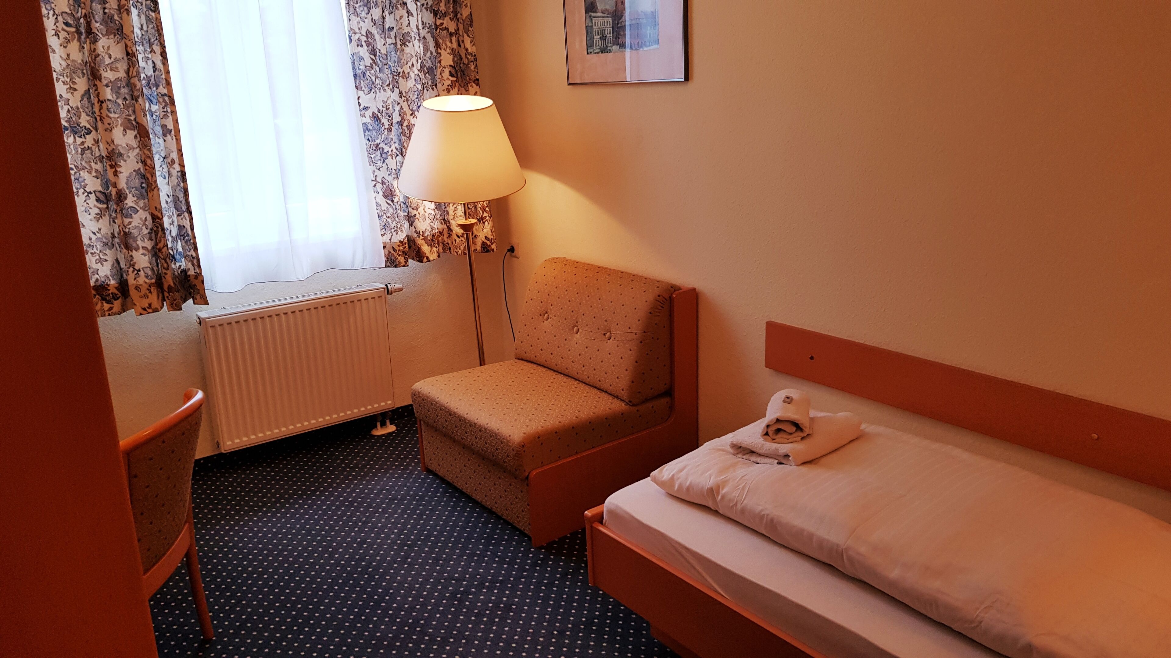 Ett hotellrum med en säng, en stol, ett skrivbord, en lampa och ett element.