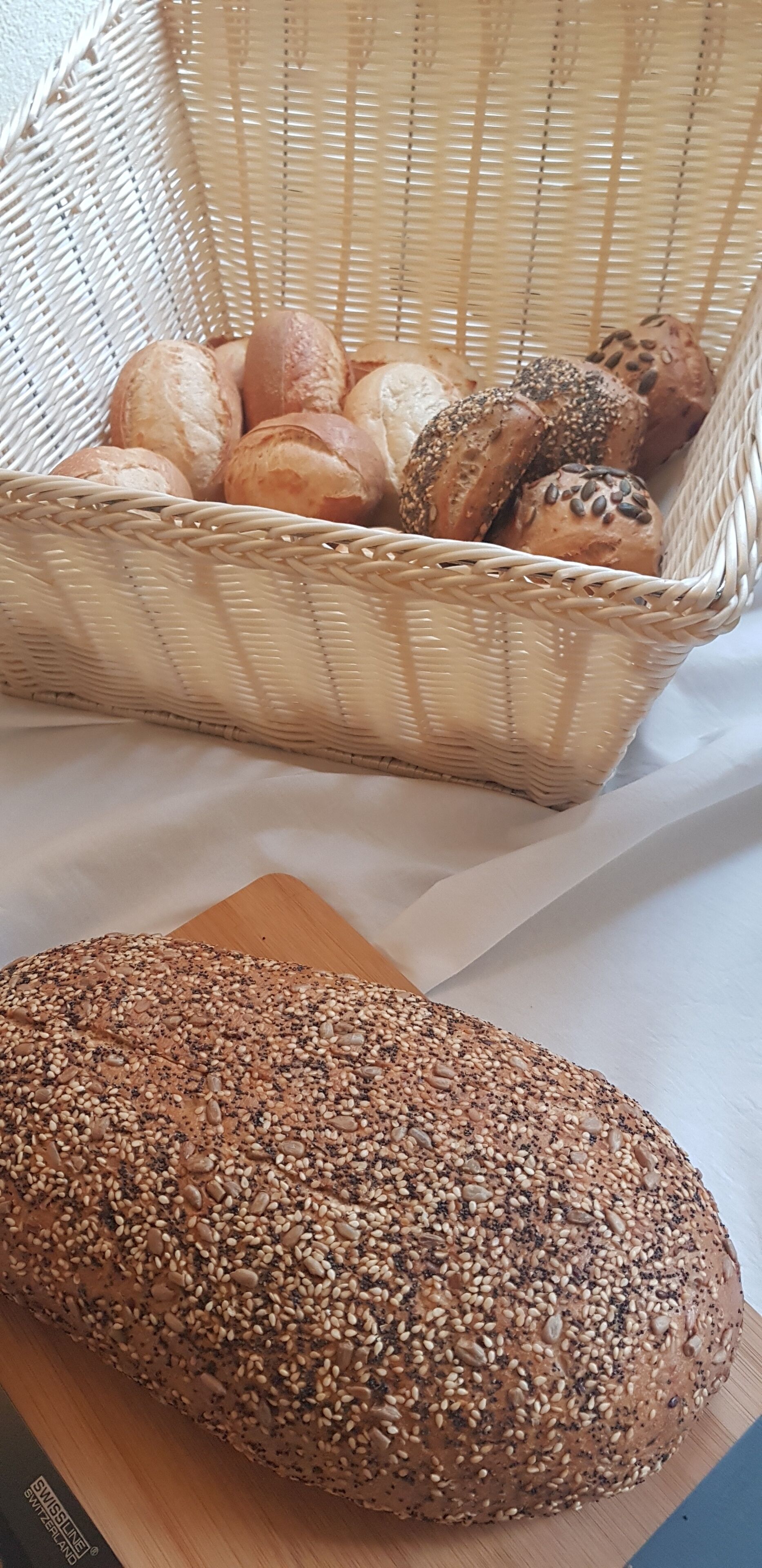 Una cesta de mimbre llena de panes variados y una hogaza de pan con semillas sobre una tabla de madera.