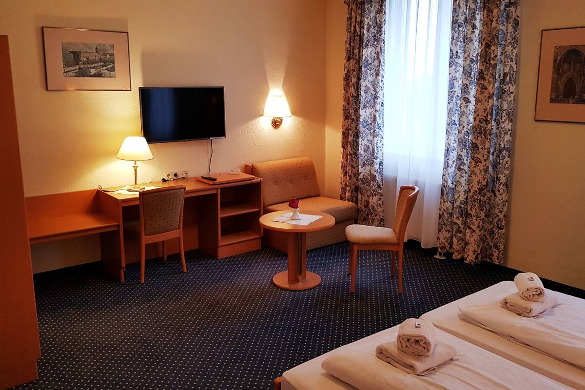 Habitación de hotel con cama, escritorio, silla, televisor y ventana con cortinas.