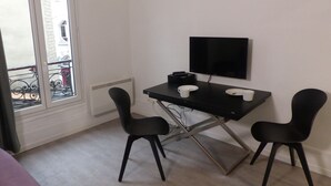 Ett litet rum med ett skrivbord, två stolar, en tv och ett fönster med utsikt över en byggnad.
