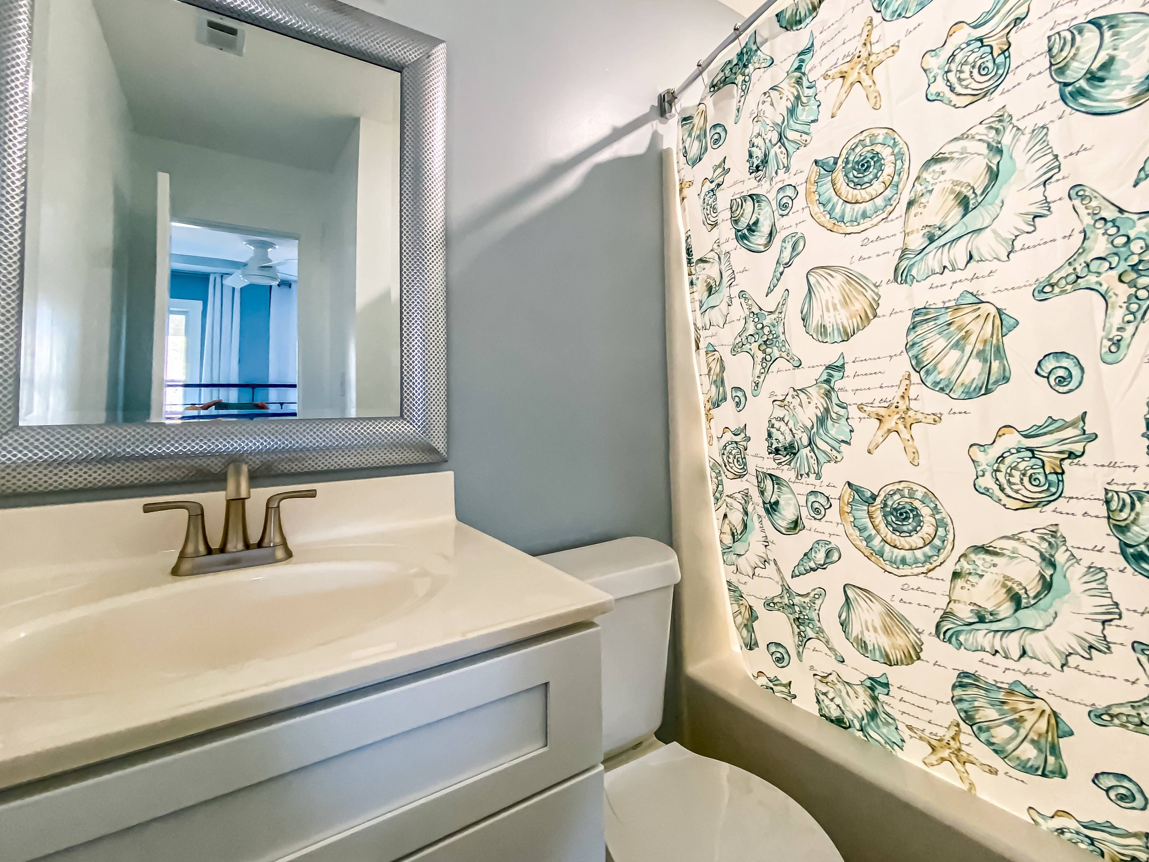 Une salle de bain avec un lavabo blanc, un miroir et un rideau de douche orné d’un motif de coquillages.