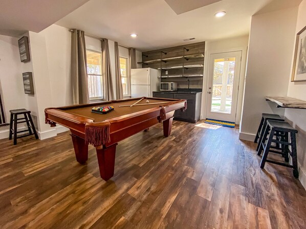 A pool table in a room with wooden flooring, a kitchen area with a microwave, and a door leading outside.