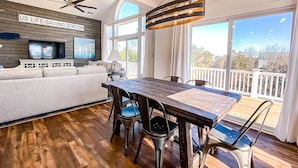 Un vaste salon doté d’un grand téléviseur, un coin repas avec une table et des chaises en bois, et un balcon offrant une vue agréable.