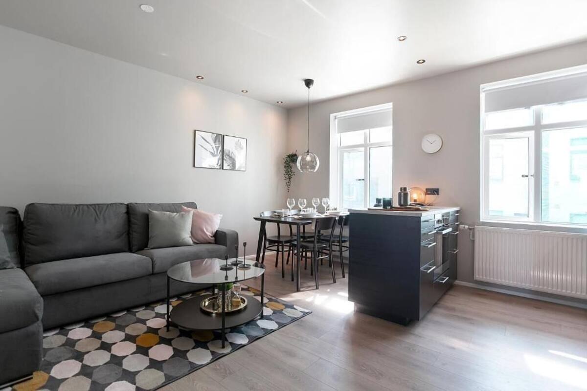 A modern living room with a grey sofa, a coffee table, and a dining area with a table and chairs. There is a kitchen area with a counter and appliances.