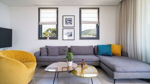 Uma sala de estar moderna com um sofá modular cinza, uma poltrona amarela e uma mesa de centro de vidro. Há duas janelas com cortinas brancas, uma TV de tela plana à esquerda e um vaso com planta no sofá.