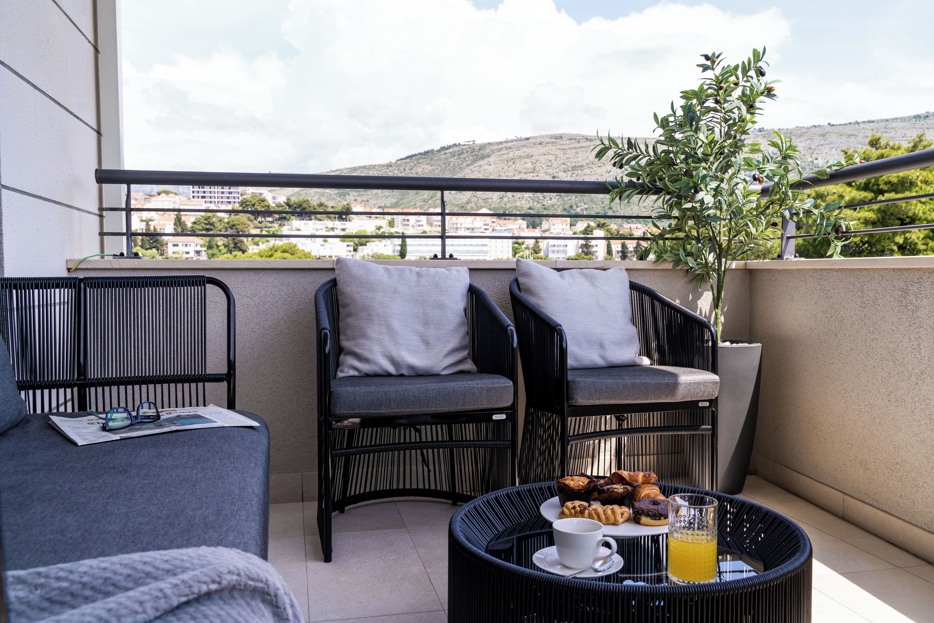 A balcony with wicker furniture, a table with pastries and a drink, and a view of a cityscape.