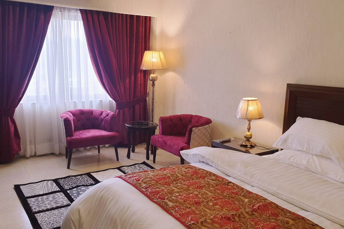 Une chambre d’hôtel avec un lit, deux fauteuils, une petite table, une lampe et une fenêtre avec des rideaux.