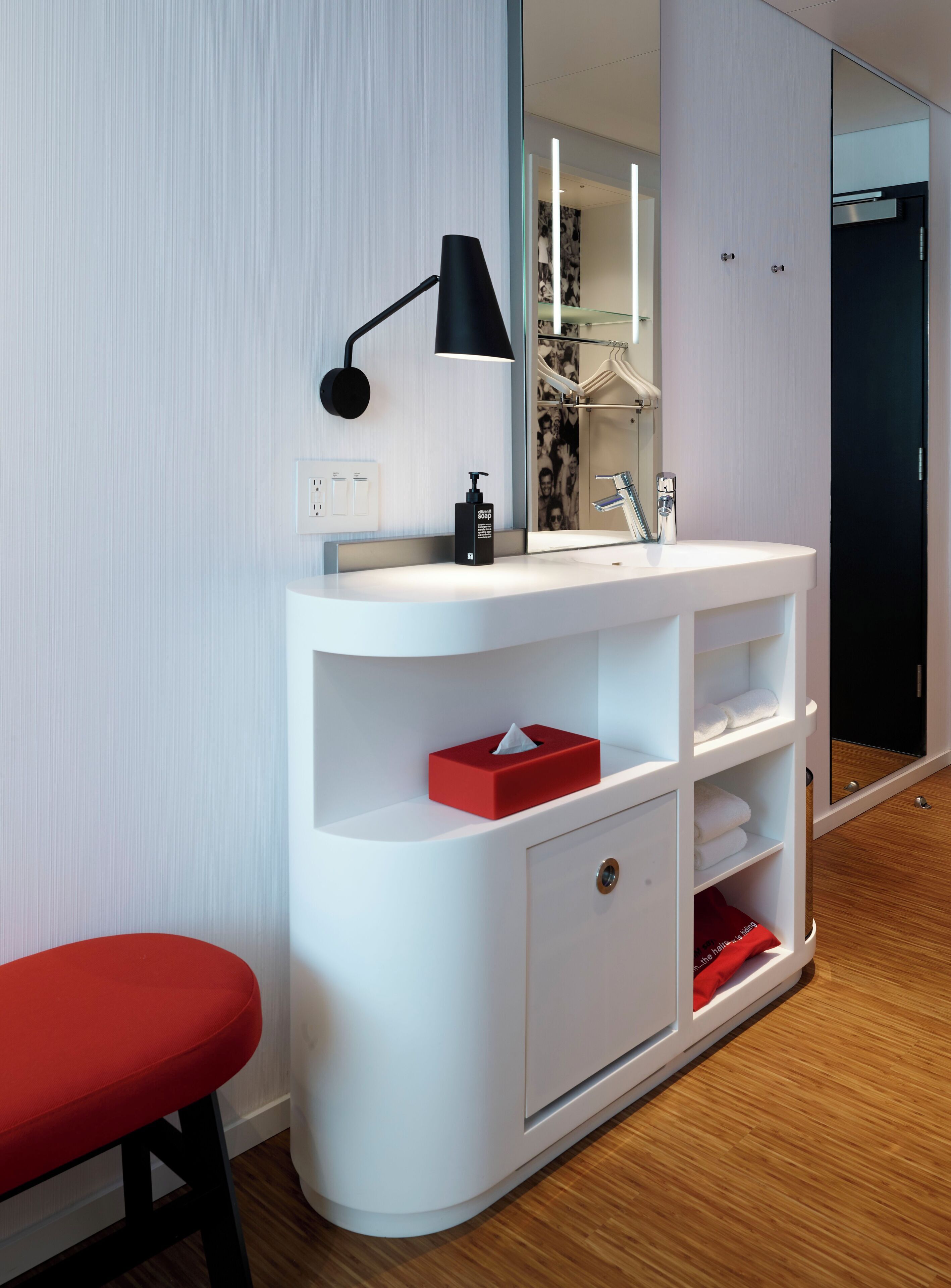A modern bathroom with a white vanity, a red stool, a black lamp, and a mirror reflecting a shower area.