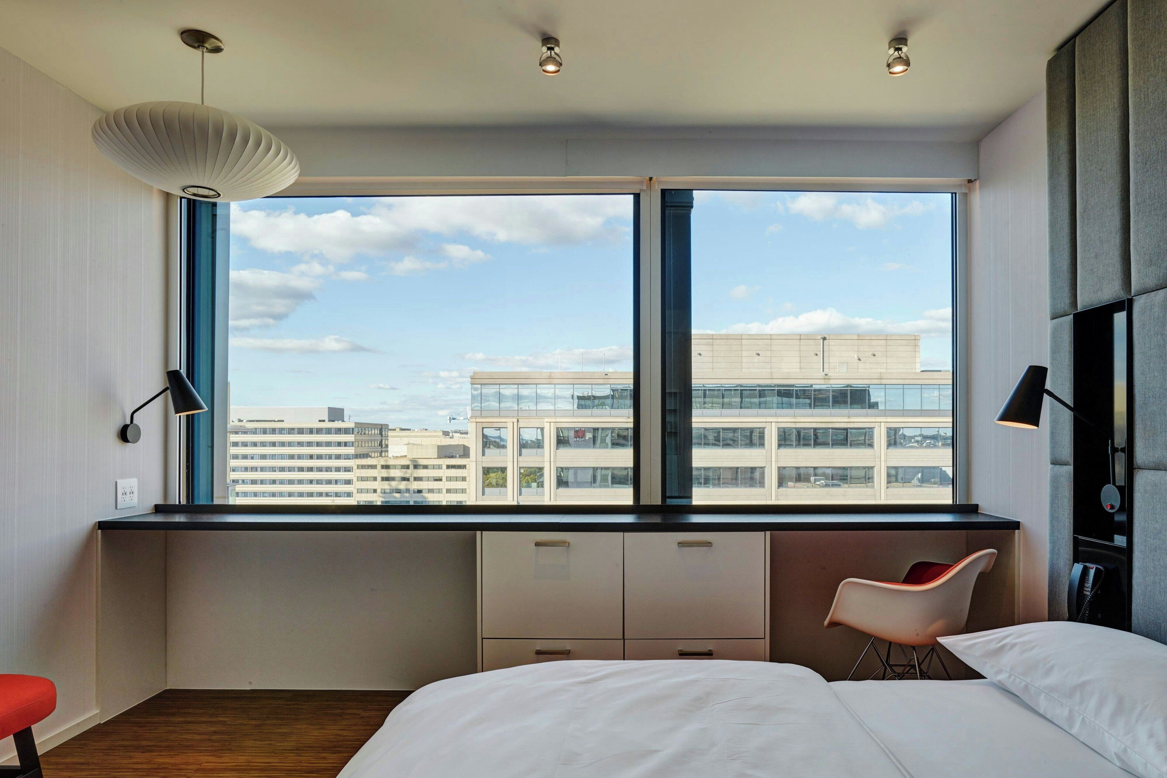 A modern hotel room with a large window, a bed, a desk, and a chair.