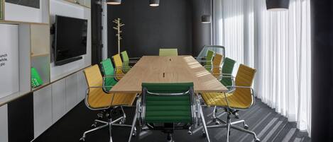 A conference room with a long table, green and yellow chairs, a TV on the wall, and a black and white striped carpet.