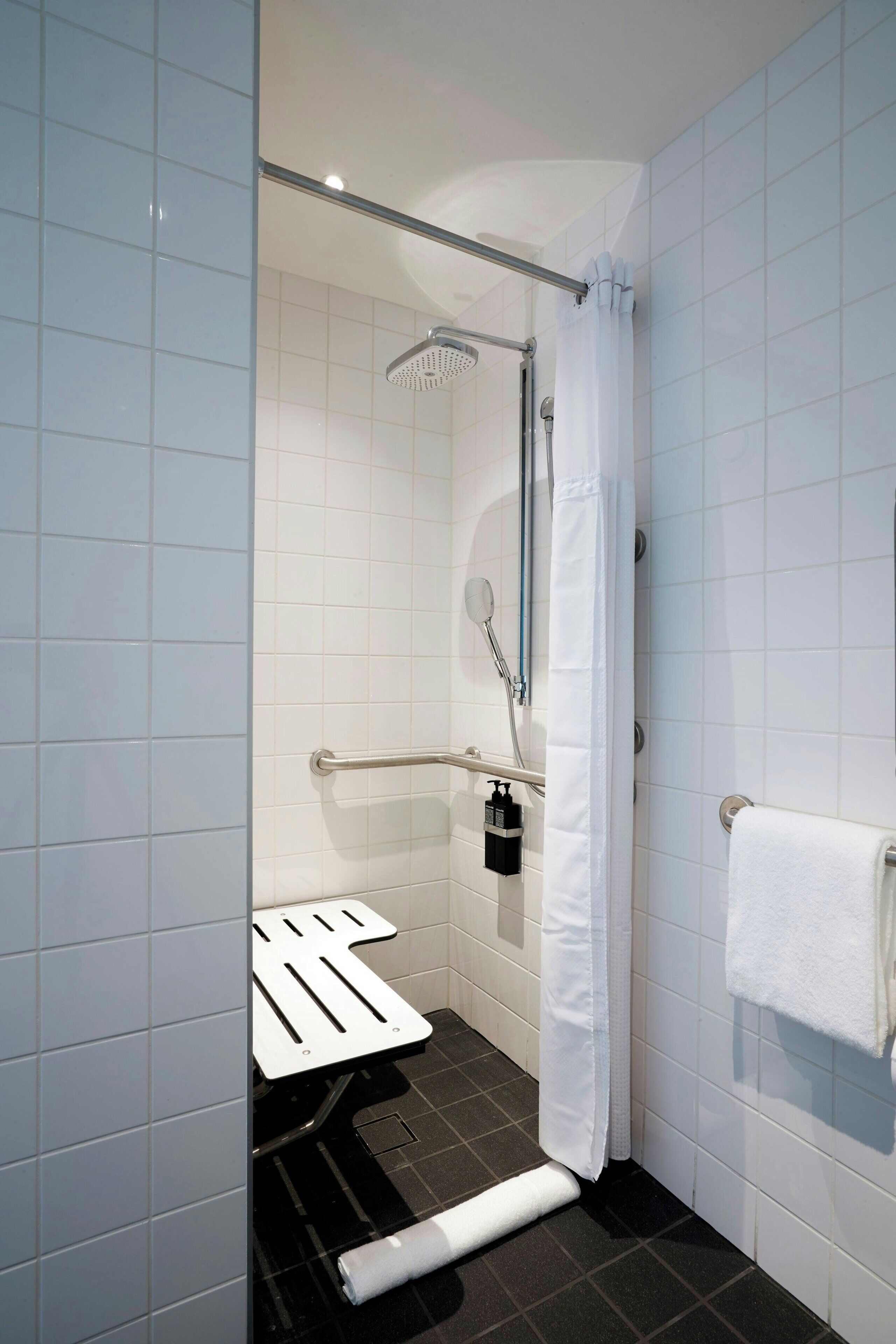 A modern bathroom with white tiles, a shower with a handheld showerhead, a grab bar, a white towel, and a black soap dispenser.