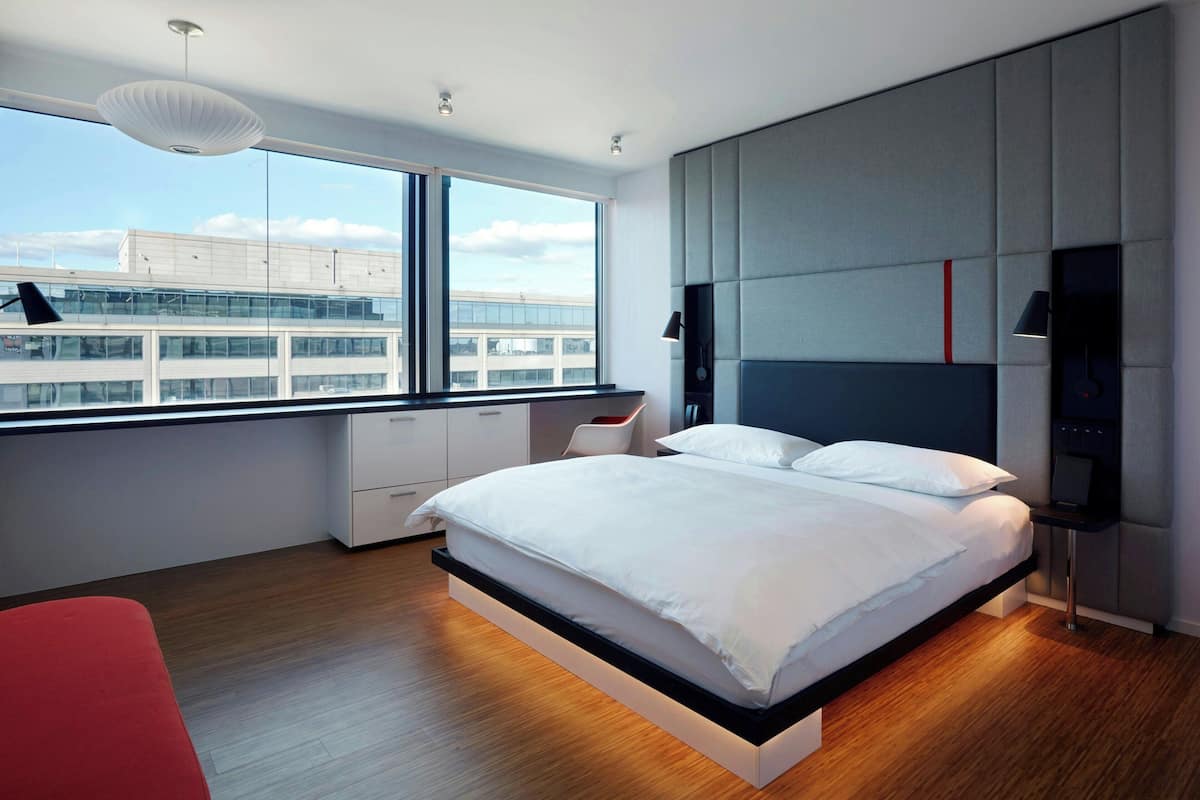 Ein modernes Schlafzimmer mit einem großen Bett, einem Schreibtisch und Blick auf eine Stadtlandschaft durch große Fenster.