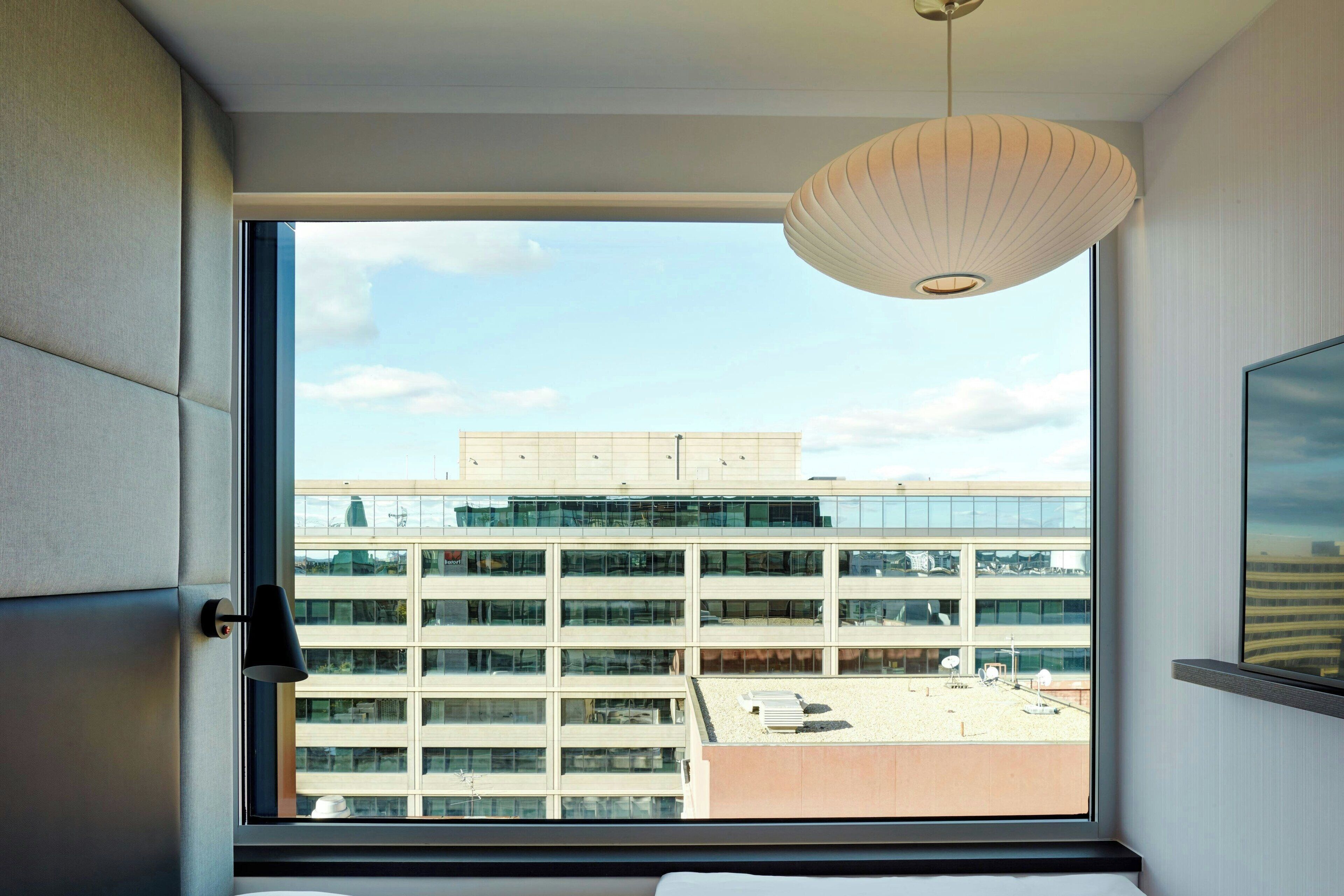 Hotellihuone, jossa on suuri ikkuna, josta avautuu näkymä moderniin rakennukseen ja kirkkaalle taivaalle.