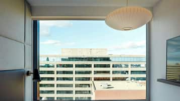 Hotellihuone, jossa on suuri ikkuna, josta avautuu näkymä moderniin rakennukseen ja kirkkaalle taivaalle.