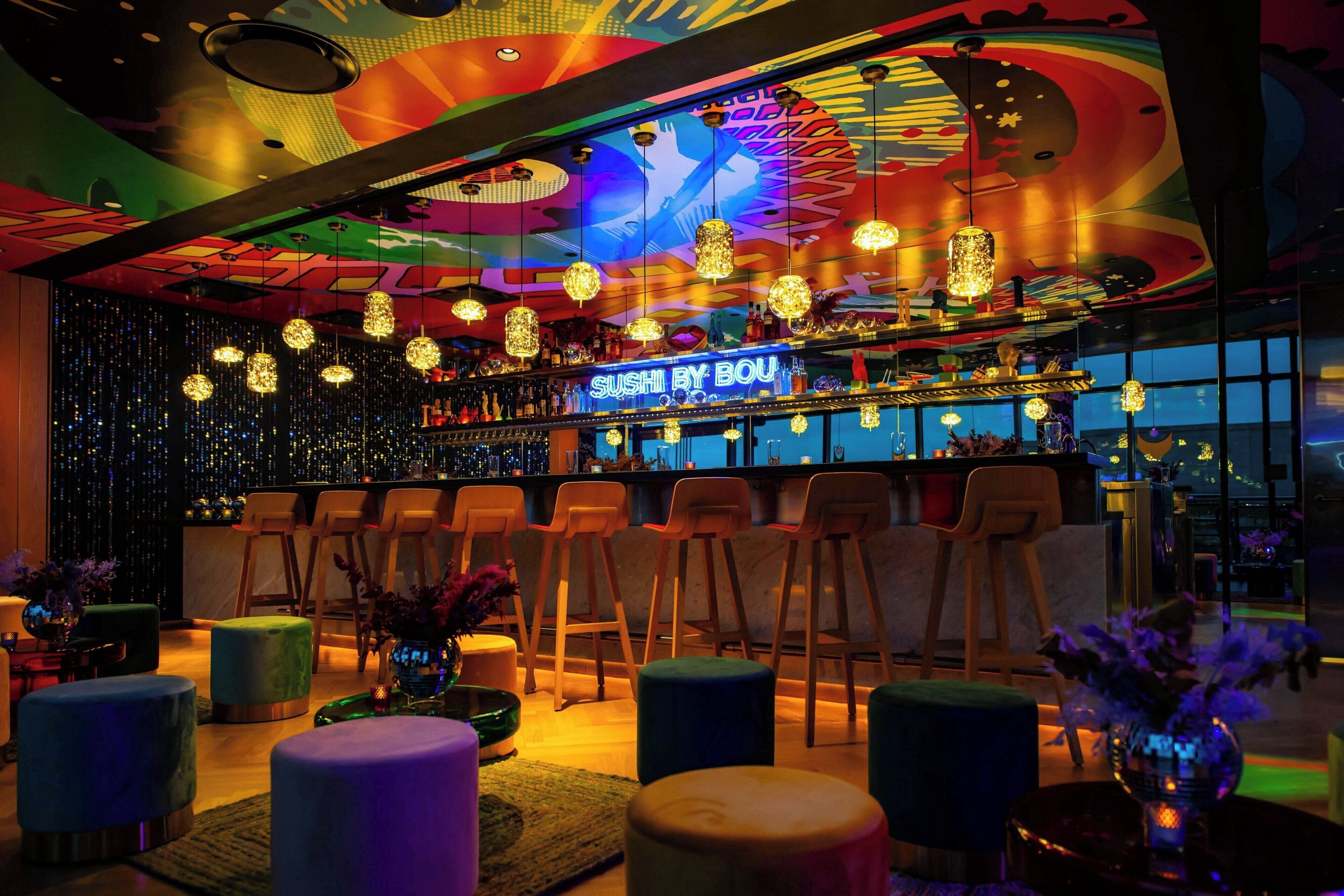 A vibrant bar area with a neon sign reading "SUSHI BY BOU", multiple hanging light fixtures, and a bar counter with stools.