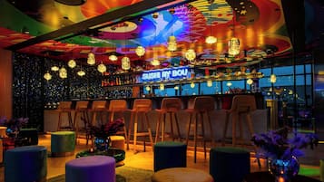 A vibrant bar area with a neon sign reading "SUSHI BY BOU", multiple hanging light fixtures, and a bar counter with stools.