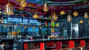 Eine Bar mit Neon-Schild, das "SUSHI BY BOU" zeigt, roten Stühlen und hängenden, kugelförmigen Leuchten.