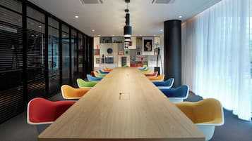 Un salón de conferencias con una larga mesa de madera y sillas coloridas, amplias ventanas con persianas y un diseño moderno de techo.