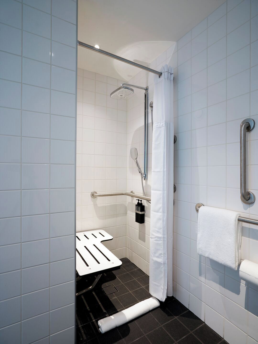 A modern bathroom with white tiles, a shower with a handheld showerhead, a grab bar, a white towel, and a toilet with a safety bar.