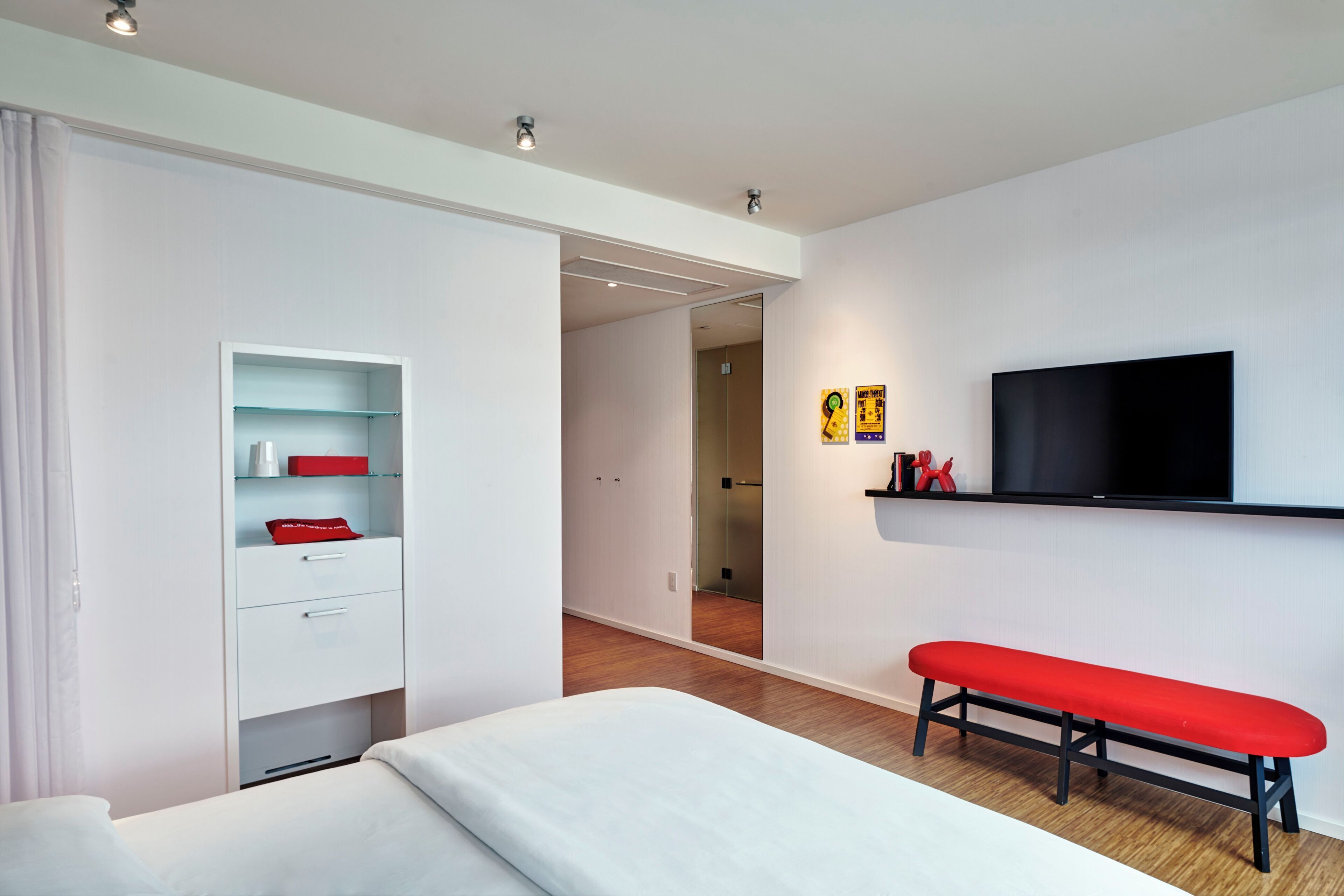 A modern hotel room with a bed, a bench, a TV, a wardrobe, and a mirror.