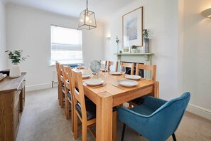 Un comedor con una mesa de madera dispuesta para cuatro personas, rodeada de sillas, un sillón tapizado en azul y una lámpara colgante.