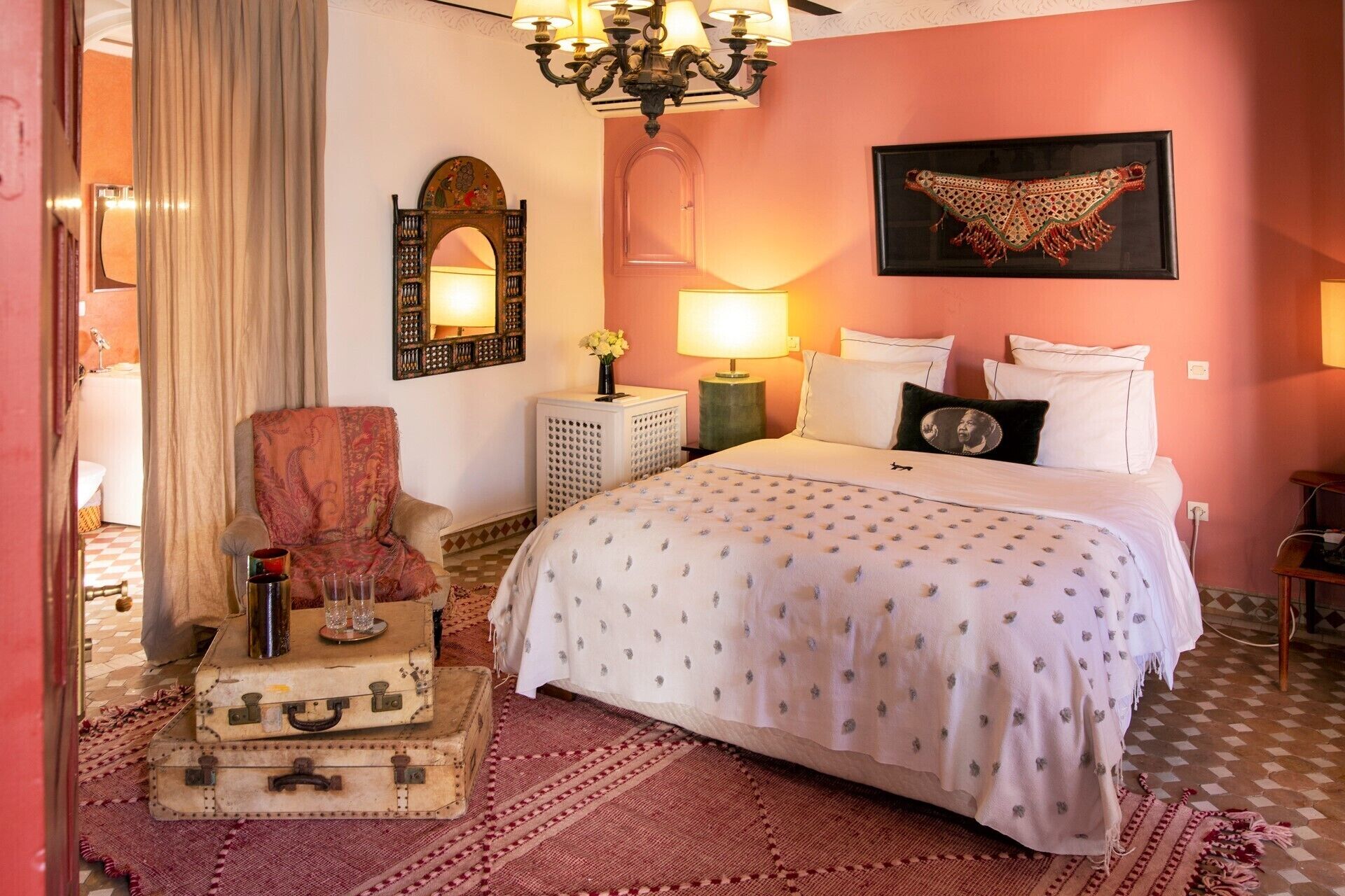 A bedroom with a bed, a chair, a lamp, a mirror, and a wall-mounted artwork.