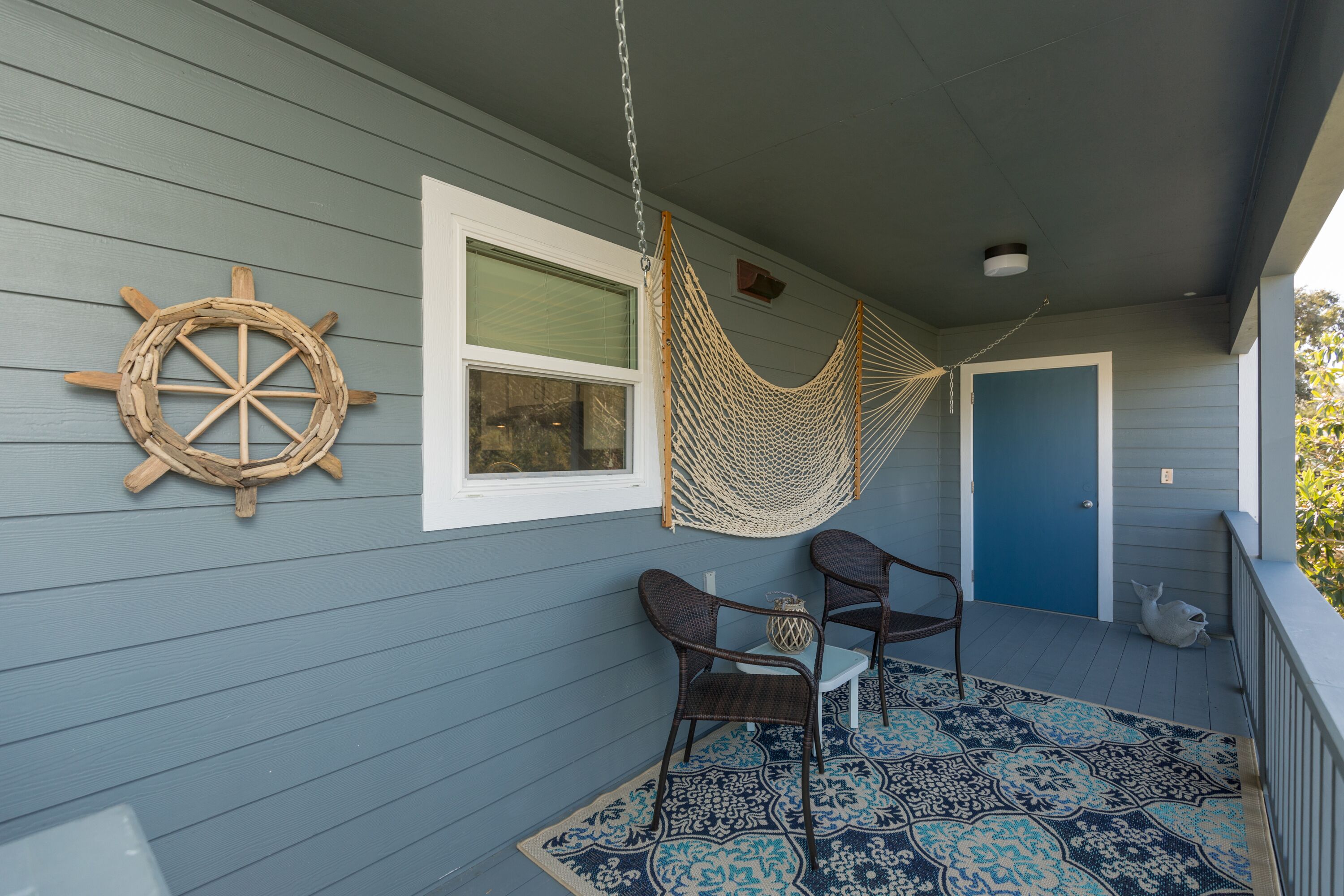 A porch with a hammock, two wicker chairs, a ship's wheel decoration, and a patterned rug.