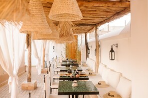 Un restaurant doté d’une terrasse, de lampes en osier et d’un toit de chaume. Les tables sont dressées avec des assiettes et des verres, et des plantes décorent l’arrière-plan.