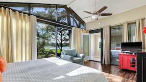 A bedroom with a large bed, a chair, a TV, and a red cabinet. There is a sliding glass door leading to a balcony with a view of trees and a lake. - Spacious 3-story home w/ stunning golf & lagoon views and large private pool (Hilton Head Island)