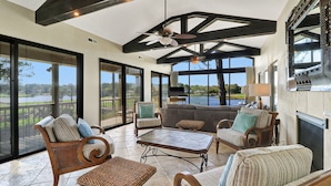 A spacious indoor area with a high ceiling, large windows offering views of a lake, and a seating area with wicker furniture. - Spacious 3-story home w/ stunning golf & lagoon views and large private pool (Hilton Head Island)
