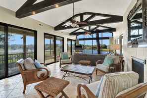 Un vaste espace intérieur doté d’un haut plafond, de grandes fenêtres offrant une vue sur un lac, et d’un coin salon avec des meubles en osier.