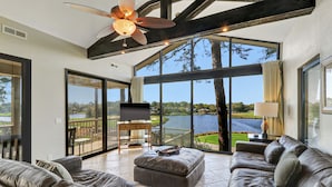 A spacious living room with a large window overlooking a lake, leather sofas, a flat-screen TV, and a ceiling fan. - Spacious 3-story home w/ stunning golf & lagoon views and large private pool (Hilton Head Island)