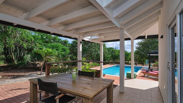 A covered patio with a wooden table and chairs, a pool in the background, and lush greenery surrounding the area.