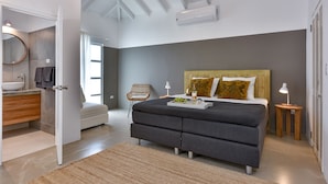 A modern bedroom with a large bed, a bedside table, a nightstand, a window, and a bathroom with a sink and mirror.