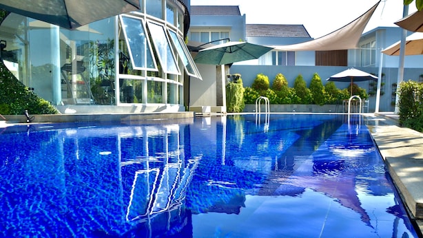 A swimming pool with clear blue water, surrounded by modern buildings and shaded by large umbrellas.