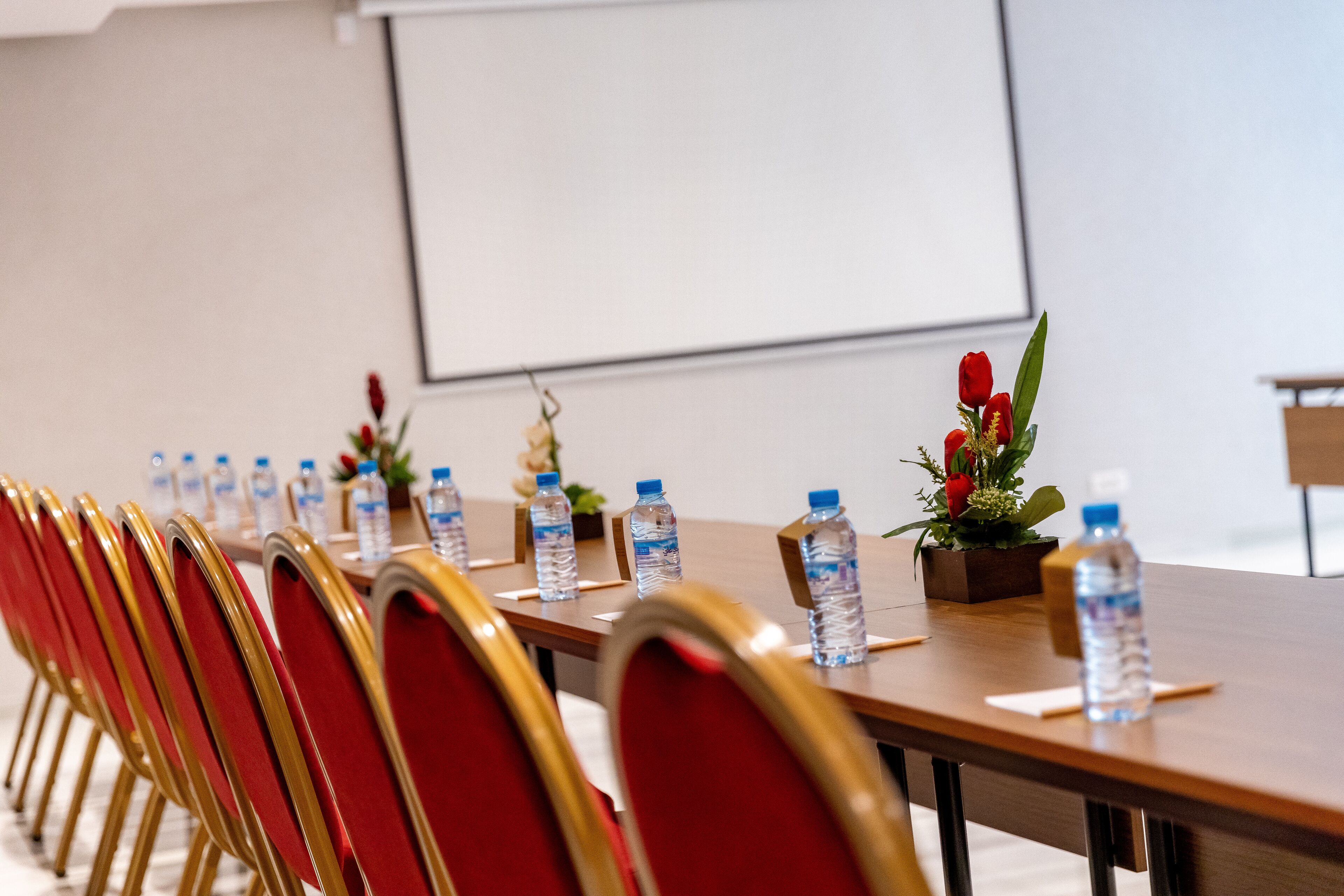 Un salón de conferencias con una larga mesa, sillas rojas, botellas de agua y una pantalla para proyector.