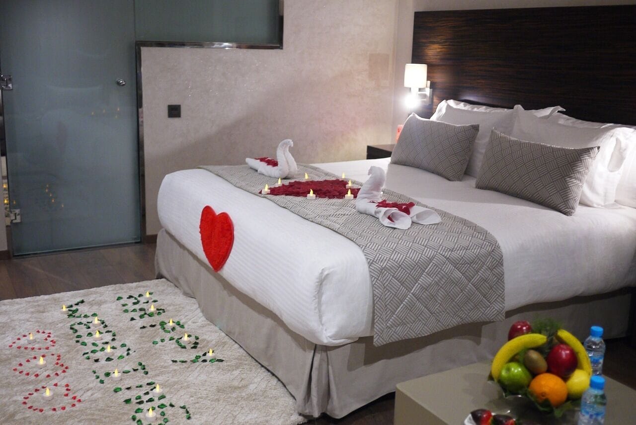 A hotel room with a large bed, a bedside table with a lamp, a fruit bowl, and a decorative rug.