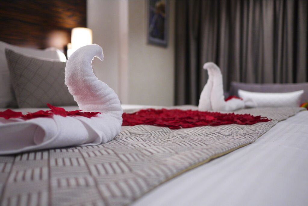 Un lit bien fait, avec une serviette blanche pliée au pied du lit, des pétales de rose rouge éparpillés sur la couverture, et un coussin à motifs.